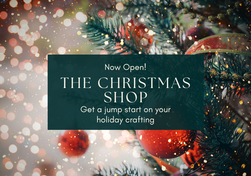 Promotional graphic for 'The Christmas Shop' with a dark teal text box reading 'Now Open! THE CHRISTMAS SHOP Get a jump start on your holiday crafting' over a festive Christmas tree background with red ornaments and gold bokeh lights.
